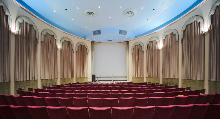 An empty recital hall with red seats and a small stage